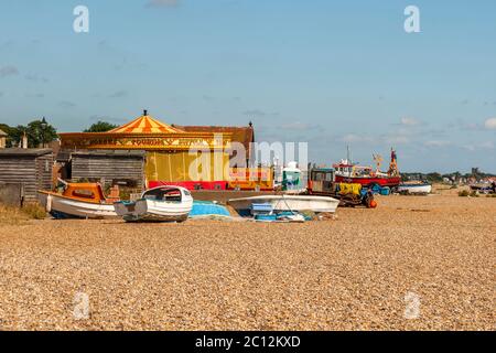 Una giostra ferma all'interno di una tenda da fiera colorata si erge sulla spiaggia di ciottoli di Aldeburgh, East Suffolk, Inghilterra; diverse piccole barche giacciono davanti ad essa, con più grandi pescherecci ormeggiati sullo sfondo. Foto Stock