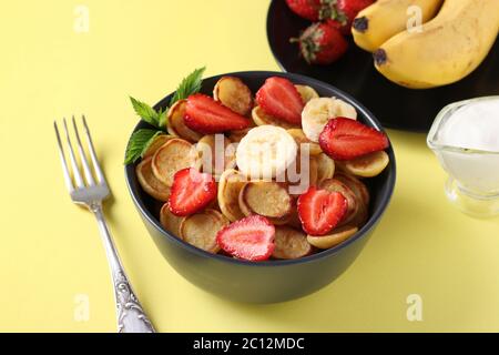 Minuscoli pancake alla moda per colazione con fragole, banana e panna acida in ciotola scura su sfondo giallo. Primo piano Foto Stock