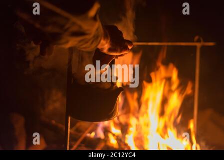 Mano di uomo che prende il bollitore, che ha riscaldato su un fuoco luminoso alla notte all'aperto Foto Stock