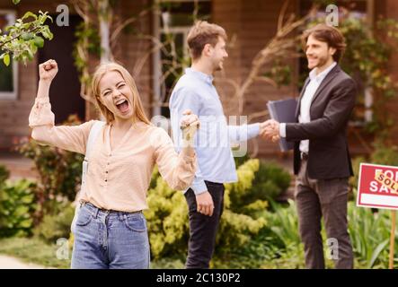 Donna millenaria estatica con chiave di casa e il suo ragazzo scuotendo le mani con broker immobiliare in cortile Foto Stock