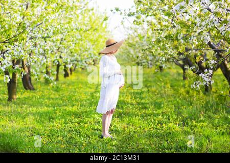 Donna incinta che indossa un cappello grande e un abito bianco in giardino fiorito Foto Stock