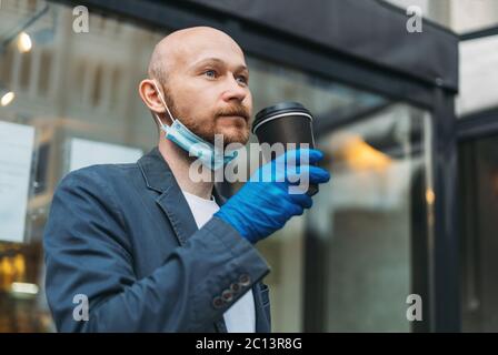 Uomo calvo adulto con la maschera medica e guanti di gomma con tazza di carta di caffè per andare. Nuovo normale, concetto di quarantena del coronavirus Foto Stock
