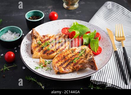 Bistecche di maiale alla griglia e insalata con pomodori in piatto su sfondo scuro. Foto Stock