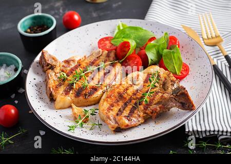 Bistecche di maiale alla griglia e insalata con pomodori in piatto su sfondo scuro. Foto Stock