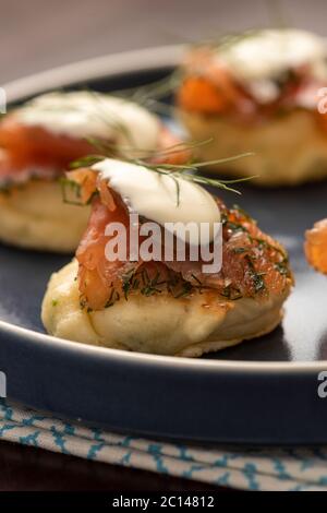 gravlax di salmone affumicato essiccato con aneto e panna acida su pancake blini e piatto di ceramica blu e tovagliolo su tavolo di legno Foto Stock