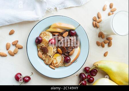 minuscoli pancake con pere, ciliegie, noci in piastra metallica sullo sfondo della tavola da cucina con frutta e latte. cibo di tendenza cereali frittelle. br Foto Stock
