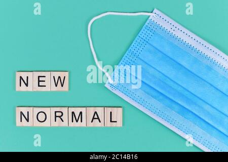 Piatto con maschera protettiva monouso blu e lettere in legno che formano "nuovo normale" su sfondo teale Foto Stock