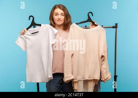 Confuso incerto donna acquirente tenere vestiti su scaffali e cercando con dubbi, pensando e scegliendo abbigliamento alla moda, shopping in boutique moderna, CAN` Foto Stock