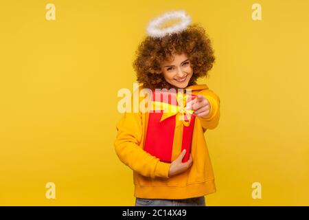 Ciao, buone vacanze! Ritratto di allegra donna angelica riccamente-capelli con san nimbus che punta alla macchina fotografica e che tiene avvolto scatola regalo di natale. Foto Stock