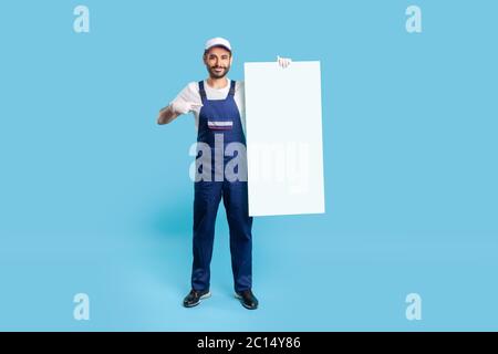 L'intera lunghezza dell'uomo da lavoro felice in guanti uniformi e protettivi che puntano alla sagoma verticale del poster, oscura lo spazio vuoto per il messaggio pubblicitario, Foto Stock