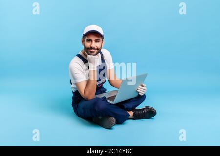 Ritratto di un simpatico e felice handyman professionista in abbigliamento da lavoro seduto a croce con il laptop, appoggiato a mano e guardando la fotocamera con amichevole smi Foto Stock