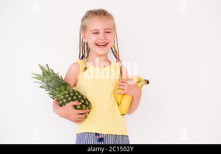 Sorriante beatiful bambina con capelli intrecciati che tengono banana e ananas su sfondo bianco Foto Stock