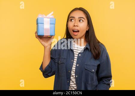 Ritratto di divertente ragazza stupita in camicia denim che tiene in mano il regalo avvolto, guardando in su con bocca aperta e l'espressione scioccata, pensando che cosa è all'interno della scatola. indo Foto Stock