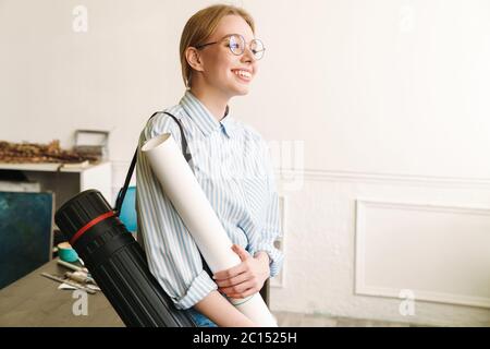 Foto di felice architetto bionda con tubo sorridente e tenendo disegno mentre si è in piedi sul posto di lavoro Foto Stock