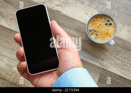 Smartphone in mano al lavoro con un'area vuota per inserire un'immagine personalizzata. Tazza di tè e pavimento in legno sullo sfondo Foto Stock