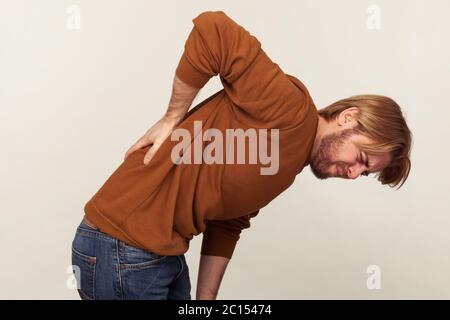 Vista laterale di un giovane sfinito malsano che cova e tocca la schiena dolorante, che soffre di disagio lombare inferiore, dolore muscolare di lavoro eccessivo, ferita spin Foto Stock