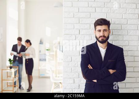 Positivo felice uomo d'affari barbuto in una giacca sorridente mentre in piedi contro uno sfondo bianco ufficio. Foto Stock