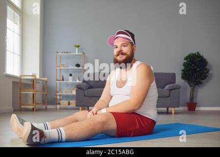 Divertente uomo grasso rosso facendo esercizi sul pavimento sorridendo mentre si siede sul pavimento a casa. Foto Stock