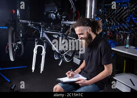 Tecnico dell'officina della bicicletta che svolge lavori amministrativi. Il meccanico della bicicletta scrive nel suo notebook di lista di controllo. Meccanico di biciclette al lavoro. Lavoratore caucasico Foto Stock