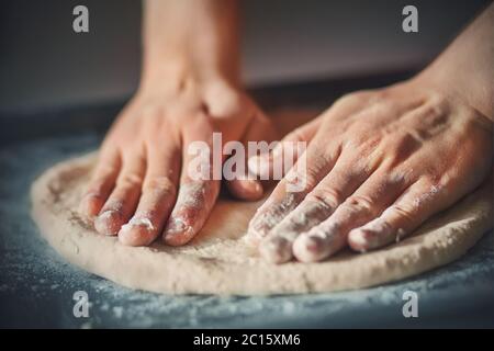 Un uomo con le mani rotola fuori la pasta fatta in casa della pizza, sdraiato su una teglia scura e illuminato dalla luce. Cucinare a casa. Foto Stock