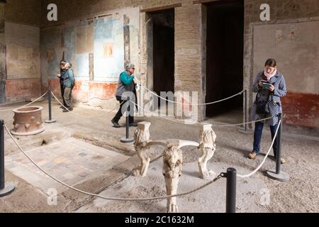 I turisti esplorano la casa di Casca Longus con pareti affrescate dipinte e un tavolo con tre supporti in marmo a forma di zampe di leone. Pompei, Italia Foto Stock