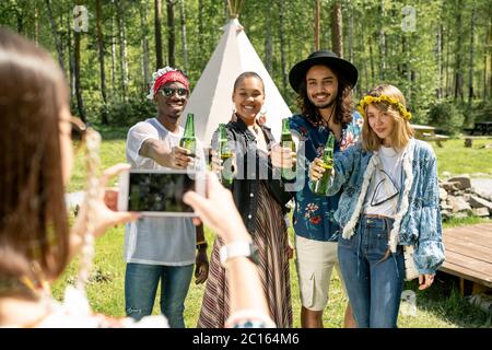 Gruppo di giovani multietnici positivi in abiti hippie che si esibiscono con bottiglie di birra per scattare foto al festival di campagna Foto Stock