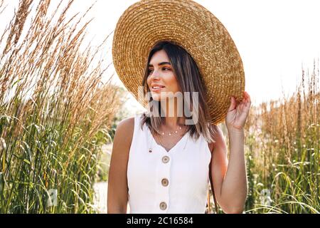 Giovane donna sorridente che indossa un cappello di paglia e cammina nel parco Foto Stock