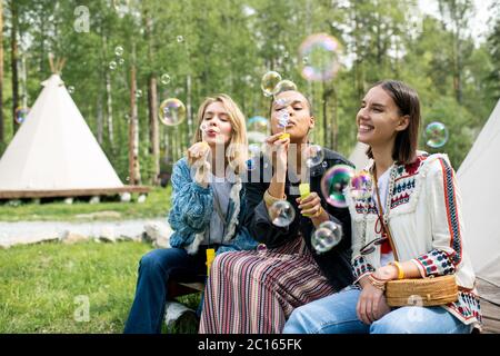 Giovani donne multietniche positive che si siedono in tenda nella foresta e soffiano bolle di sapone Foto Stock