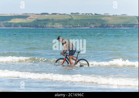 Waterford, Irlanda. 14 Giugno 2020. Tramore Beach era occupato oggi in una giornata gloriosamente soleggiata con alti da 18 a 23 gradi Celsius. Un uomo guida la sua bici nel mare per rinfrescarsi. Credit: Notizie dal vivo di AG/Alamy Foto Stock