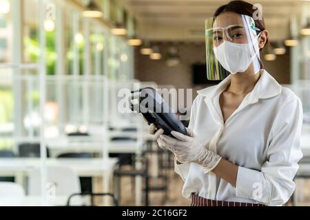 Ritratto attraente cameriera asiatica indossare maschera viso e visiera tenere lettore di carte di credito per il pagamento senza contatto con sfondo ristorante interno. N