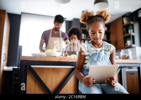 Carino African American Girl utilizzando una compressa mentre i suoi genitori nella preparazione degli alimenti in cucina Foto Stock