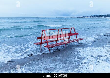 La panca rossa coperta di ghiaccio è colpita dal mare invernale. Onde ghiacciate del Mar Baltico su una spiaggia vuota. Foto Stock