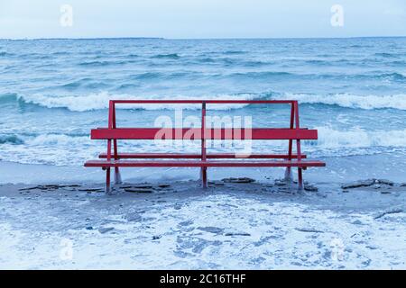 La panca rossa coperta di ghiaccio è colpita dal mare invernale. Onde ghiacciate del Mar Baltico su una spiaggia vuota. Foto Stock