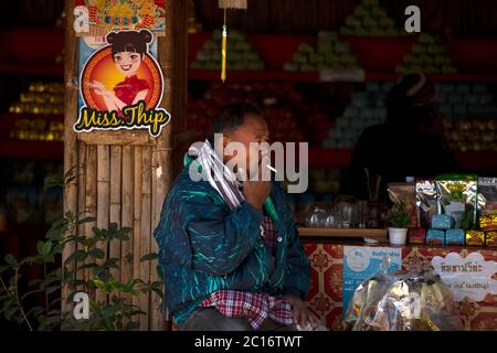 Un uomo seduto fuori da un negozio e fumando nel villaggio tailandese cinese di Ban Rak, provincia di Mae Hong Son, Thailandia del nord. Foto Stock