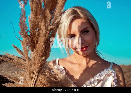 Ritratto di una bella ragazza bionda con capelli lunghi in un campo una giornata di sole al tramonto. Nelle sue mani tiene l'erba gialla alta e asciutta. Campagna. Foto Stock