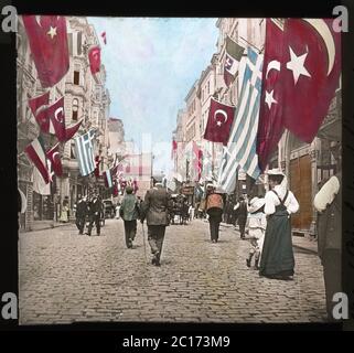 Viale İstiklal o Via İstiklal (in turco: İstiklal Caddesi; in francese: Grande Rue de Péra; in inglese: Viale dell'indipendenza) è uno dei viali più famosi di Istanbul, Turchia. Durante il periodo ottomano, il viale è stato chiamato Cadde-i Kebir (Grand Avenue) in turco o Grande Rue de Péra. La fotografia mostra un gruppo di turisti europei che camminano per la strada durante le celebrazioni dell'anniversario dell'incoronazione del sultano. Vetrino colorato a mano da circa 1910. Fotografia su lastra di vetro asciutta della collezione Herry W. Schaefer. Foto Stock