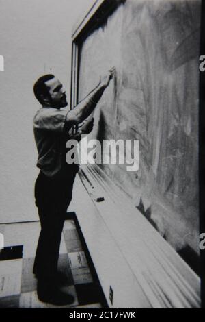Fine anni 70 vintage in bianco e nero fotografia estrema di un insegnante in piedi alla lavagna e di scrittura su di esso. Foto Stock