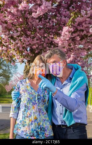 Affettuosa coppia senior che indossa maschere e guanti colorati durante il braccio in braccio pandemico coronavirus che ha una discussione e un puntamento Foto Stock