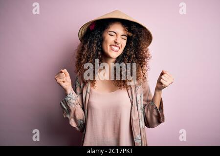 Giovane bella donna con capelli ricci e piercing con cappello conico asiatico tradizionale molto felice ed eccitato facendo gesto vincitore con le braccia sollevate, Foto Stock