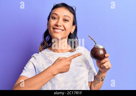 Giovane donna bevendo l'infusione di compagno sorridente felice indicando con mano e dito Foto Stock