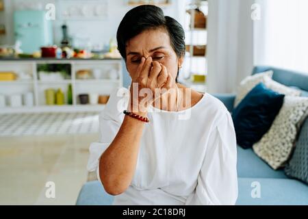 Signora asiatica matura con occhi chiusi sfregando il naso mentre si siede sul divano e soffre di mal di testa in un accogliente soggiorno a casa Foto Stock