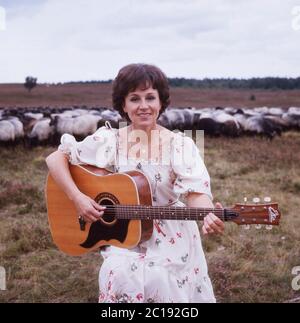 Lolita, österreichische Schlagersängerin, singt mit einer Gitarre vor einer Herde Schafe in der Lüneburger Heide, Deutschland um 1984. La cantante austriaca Lolita suona una chitarra e canta con un gregge di pecore a Lueneburg Heath, Germania, intorno al 1984. Foto Stock