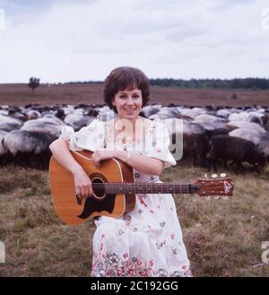 Lolita, österreichische Schlagersängerin, singt mit einer Gitarre vor einer Herde Schafe in der Lüneburger Heide, Deutschland um 1984. La cantante austriaca Lolita suona una chitarra e canta con un gregge di pecore a Lueneburg Heath, Germania, intorno al 1984. Foto Stock