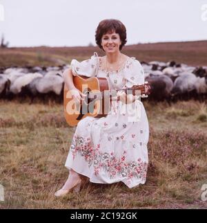 Lolita, österreichische Schlagersängerin, singt mit einer Gitarre vor einer Herde Schafe in der Lüneburger Heide, Deutschland um 1984. La cantante austriaca Lolita suona una chitarra e canta con un gregge di pecore a Lueneburg Heath, Germania, intorno al 1984. Foto Stock