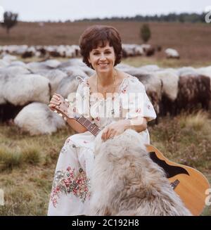 Lolita, österreichische Schlagersängerin, singt mit einer Gitarre vor einer Herde Schafe in der Lüneburger Heide, Deutschland um 1984. La cantante austriaca Lolita suona una chitarra e canta con un gregge di pecore a Lueneburg Heath, Germania, intorno al 1984. Foto Stock