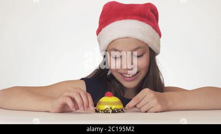 Bella ragazza teen in cappello Babbo Natale con un appetito osserva ostrfully la torta di compleanno su sfondo bianco Foto Stock