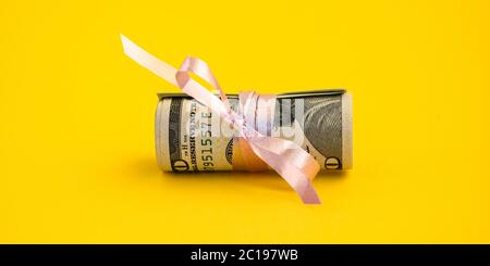 Piccolo regalo del dollaro, pacchetto delle fatture di cento dollari legati con un nastro rosa. Dollari isolati su sfondo giallo Foto Stock