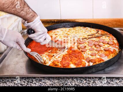 Chef affettare una pizza italiana con quattro condimenti assortiti usando un coltello e una forchetta in una cucina pizzeria da vicino Foto Stock