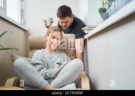 Donna bionda seduta su poltrona beige e uomo brunetto in piedi accanto a lei in appartamento Foto Stock