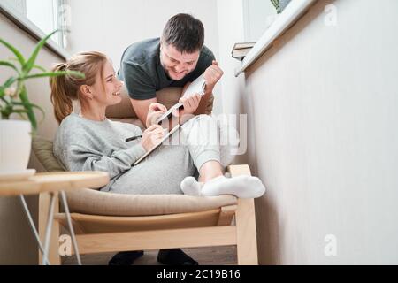 Donna sorridente seduta su poltrona beige e brunetto in piedi accanto lei sul balcone in appartamento Foto Stock